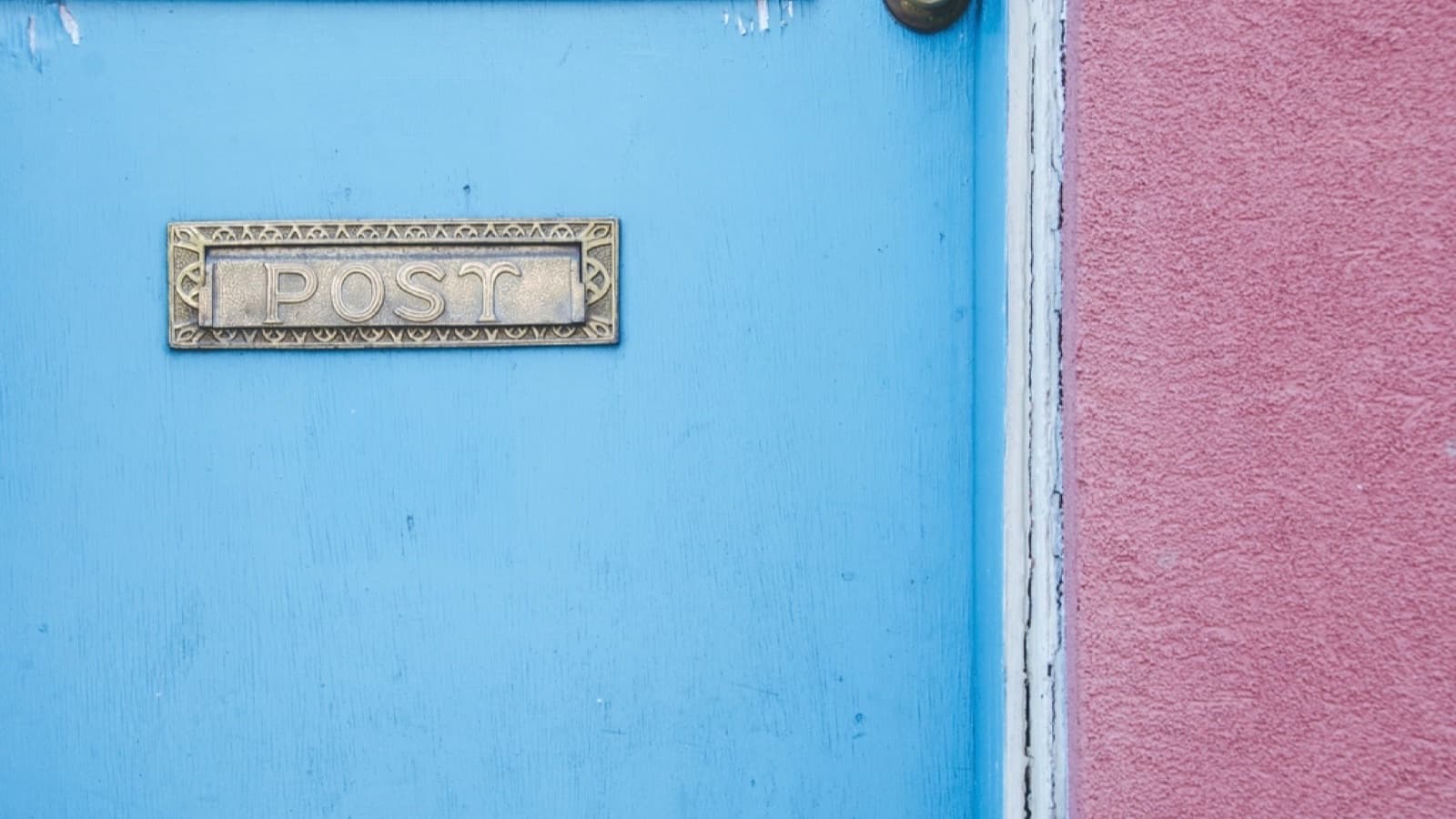 A brass letterbox slot engraved POST in a blue door beside a pink wall