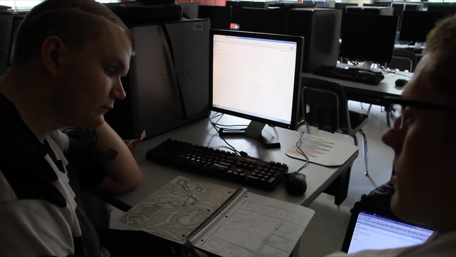 Two students at a computer in a lab, one sketching in a notebook