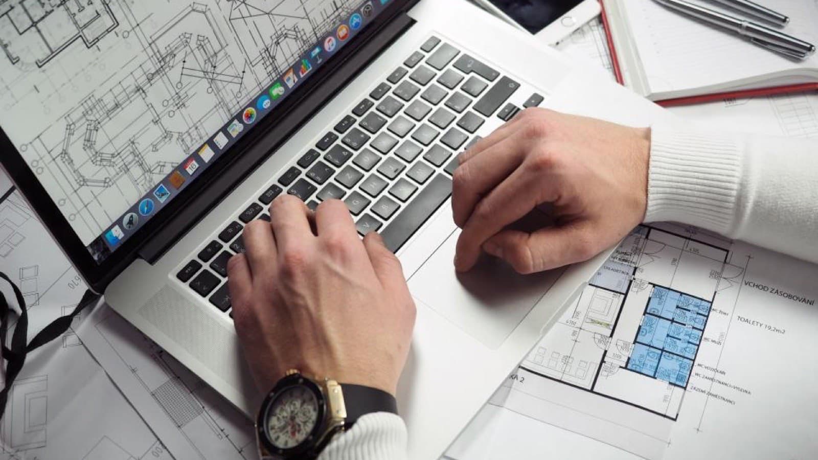 Hands typing on a laptop beside printed technical drawings on a desk