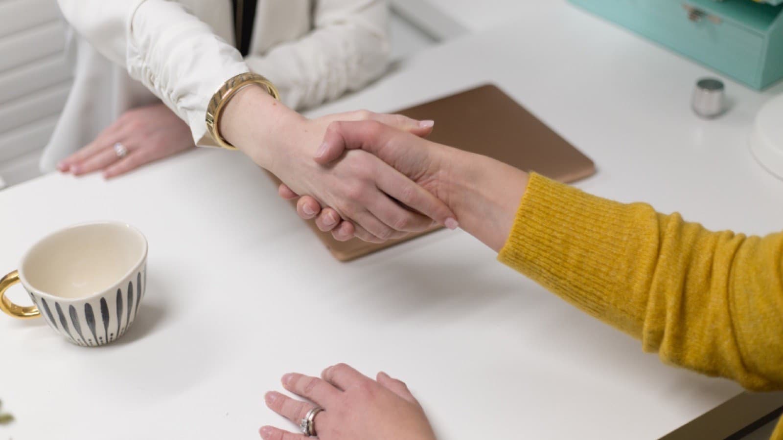 Two people shaking hands across a desk beside a coffee cup