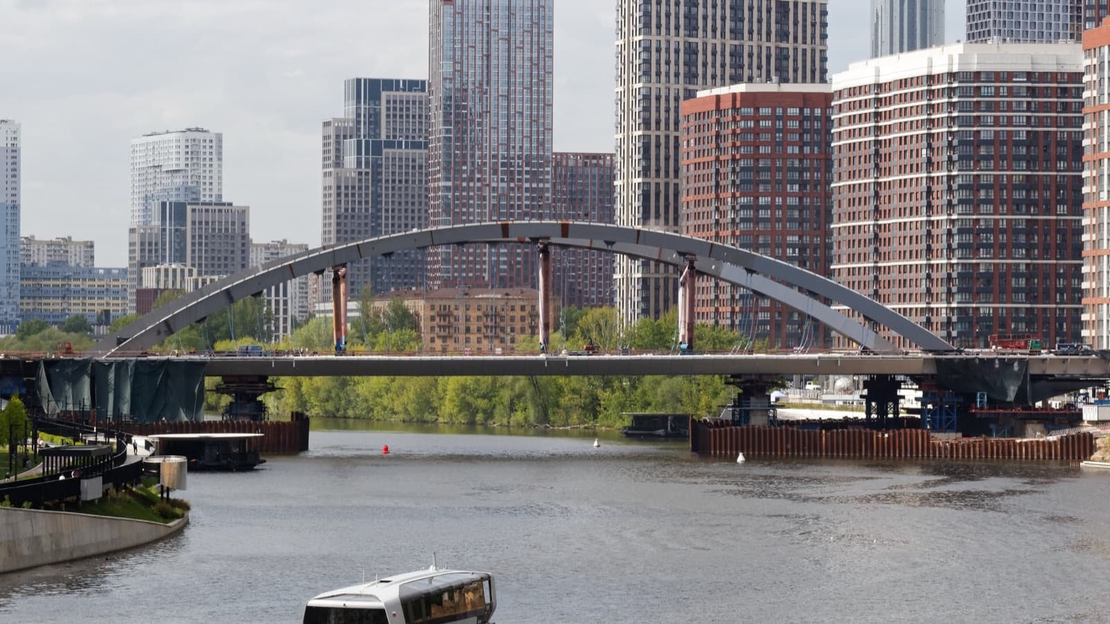 A steel arch bridge spanning a river in front of a city skyline, with a boat passing beneath