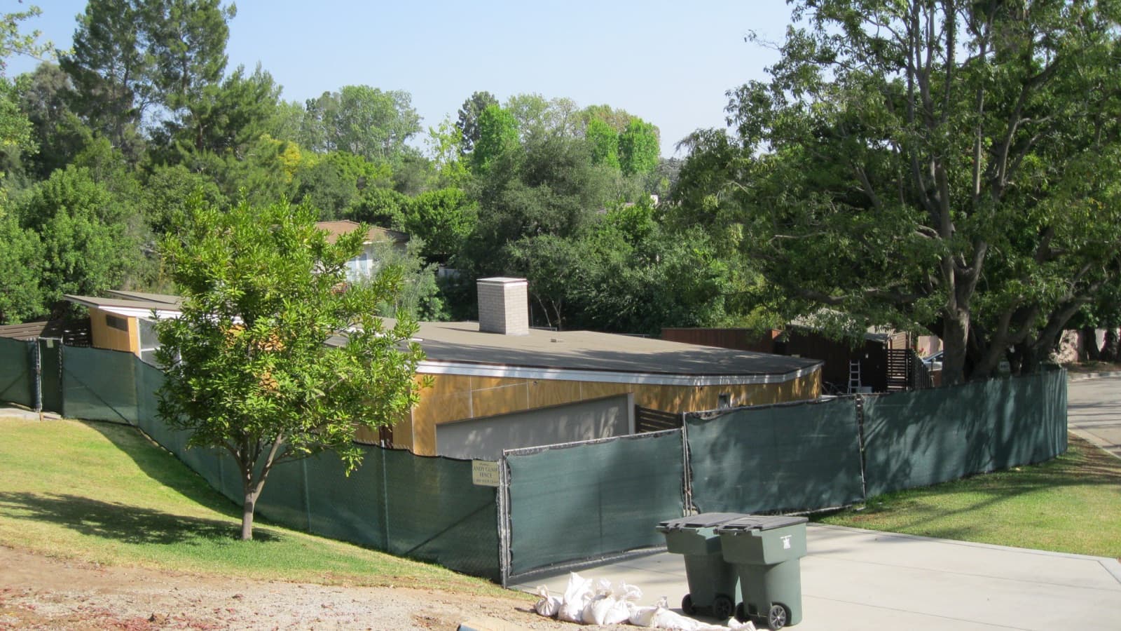 A single-storey house behind green construction fencing, with sandbags and a wheelie bin in the foreground under trees